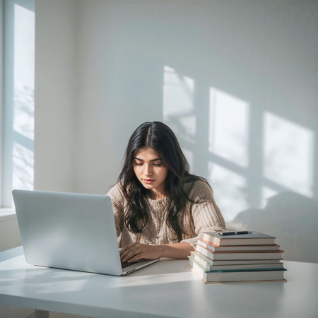 Young woman studying with a laptop and books at a desk in natural sunlightの素材