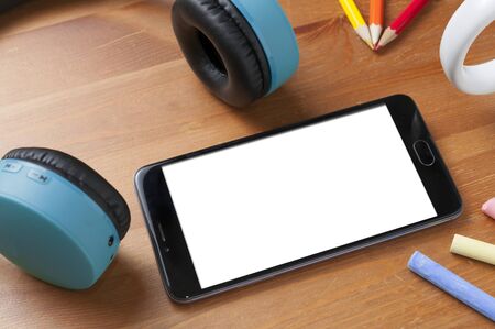 Blank screen smartphone on wooden table next to a pair of wireless earphones, some pencils and some chalksの写真素材