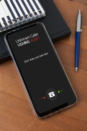 A smartphone leaning against a diary and a notepad on a wooden table show an unknower caller call with an alert and a reminder to not share personal bank dataの写真素材