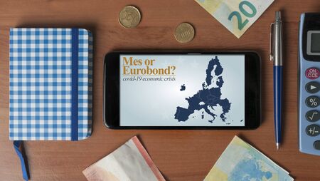 Covid-19 economic crisis: a smartphone with a question about mes or eurobond on a wooden desk with some office objectsの写真素材