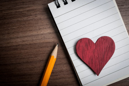 Red wooden heart on a notepad and a yellow pencil on a wooden table