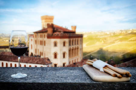 A chalice of Barolo red wine on a windowsill with breadsticks and the castle of Barolo (Piedmont, Italy) on the backgroundの写真素材