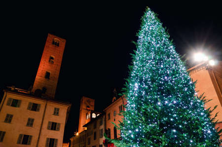 Piazza Risorgimento, main square of Alba (Piedmont, Italy) at night with the town hall, the medieval towers and an illuminated christmas treeの写真素材