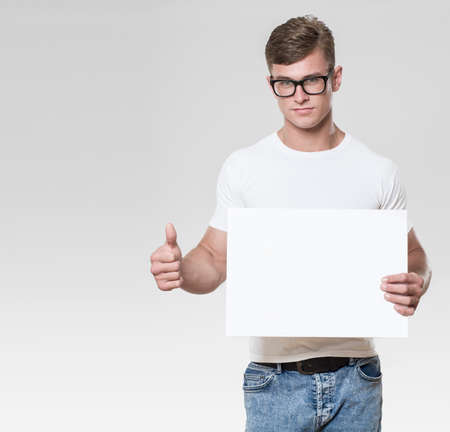 Handsome young man holding a whitebourd with thumbs up and pointing at camera. Picture taken in a studio with grey background. The model is wearing glasses and a white t-shirt.の写真素材