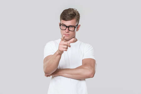 Handsome young man pointing at camera. Picture taken in a studio with grey background. The model is wearing glasses and a white t-shirt.の写真素材