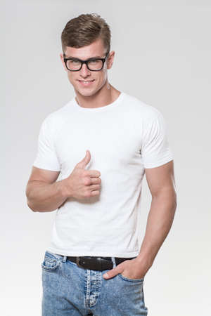 Handsome young man with thumbs up. Picture taken in a studio with grey background. The model is wearing glasses and a white t-shirt.の写真素材