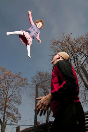 happy toddler girl jumping up on trampoline assisted by fatherの写真素材
