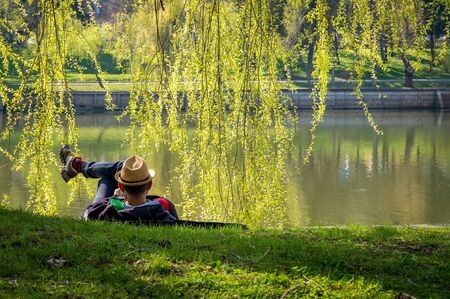 A man with a hat standing on the grass at the edge of a lake. Man enjoying a beautiful day in the park.の写真素材