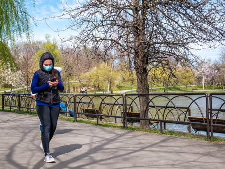 Bucharest/Romania - 03.17.2020: Woman wearing face mask to protect herself from Coronavirus. Woman walking in the park and looking at her phone.のeditorial素材