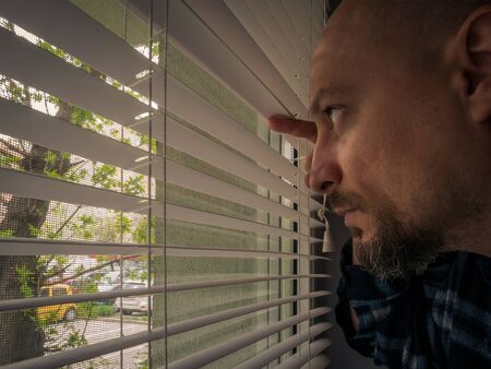 Man in isolation looking through window blinds. Man forced to stay inside the house as a result of the restrictions caused by the Coronavirus outbreak.の写真素材