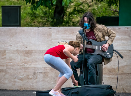 Bucharest/Romania - 05.23.2020: Young man with long hair playing guitar. Girl giving money to a street artist performing.のeditorial素材