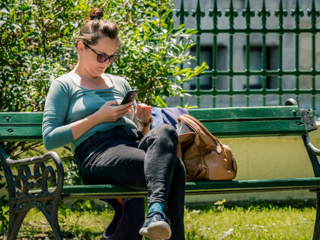 Bucharest/Romania - 05.30.2020: Woman sitting on a bench in the park and looking at her phone. Smart phone addiction conceptのeditorial素材