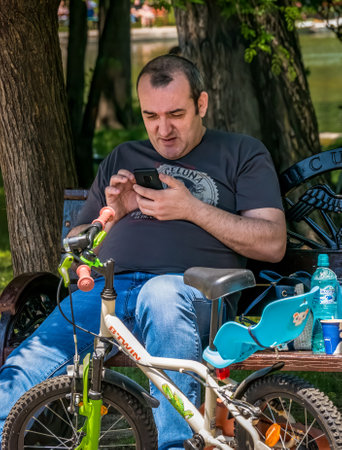 Bucharest/Romania - 05.30.2020: Man sitting on a bench in the park and looking at his phone. Smart phone addiction concept.のeditorial素材