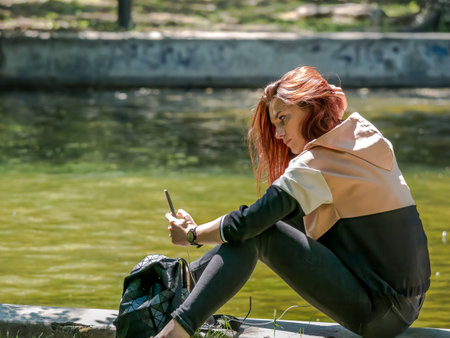 Bucharest/Romania - 05.30.2020: Girl sitting on the edge of a lake in the park and looking at her phone. Smart phone addiction concept.のeditorial素材