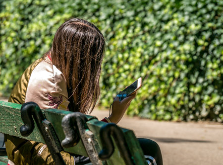 Bucharest/Romania - 05.30.2020: Woman sitting on a bench in the park and looking at her phone. Smart phone addiction concept.のeditorial素材