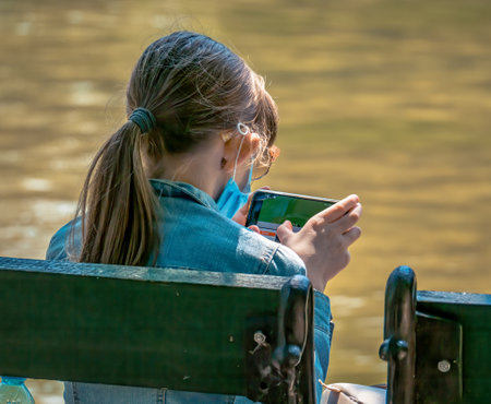 Bucharest/Romania - 05.30.2020: Young girl sitting on a bench in the park and looking at her phone. Smart phone addiction concept.のeditorial素材