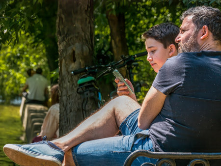 Bucharest/Romania - 05.30.2020: Young boy with his father sitting on a bench in the park and looking at her phone. Smart phone addiction concept.のeditorial素材