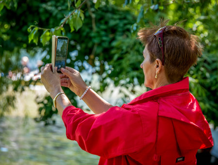 Bucharest/ Romania - 05.30.2020: Woman with a red jacket taking a picture with her phone in Cismigiu park, in Bucharest. Smartphone addiction concept.のeditorial素材