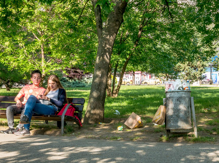 Bucharest/Romania - 05.30.2020: Garbage bin full of trash. Overflowing garbage bin and a couple sitting on bench. Environmental concept.のeditorial素材