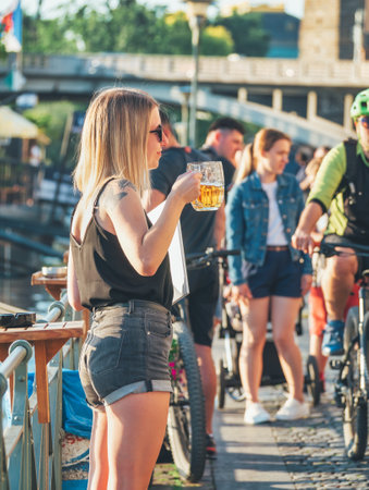 Prague, Czech Republic - June 2022: Young attractive blonde girl having a beer on Naplavka along the Vltava river, in Pragueのeditorial素材