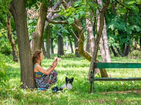 Bucharest, Romania - June 2022: Young girl sitting and relaxing on the green grass with her dog pet in the Youth park ( Tineretului)のeditorial素材