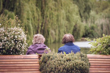 Rear view with two unidentified senior women sitting on a bench in the park with a lake in the background.の写真素材