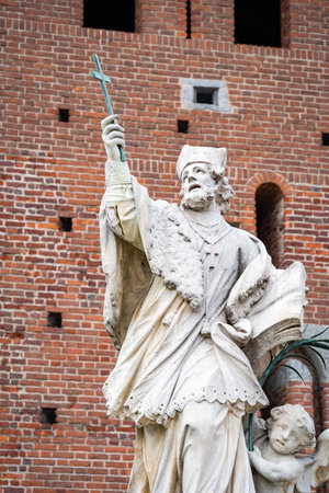 Marble statue of a saint holding a cross in front of the historic brick walls of Castello Sforzesco in Milan, Italyの写真素材