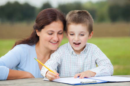 Happy little young boy in white shirt and his smiling mother write or draw with a pencil on a sheet of paper on wood table in the parkの写真素材