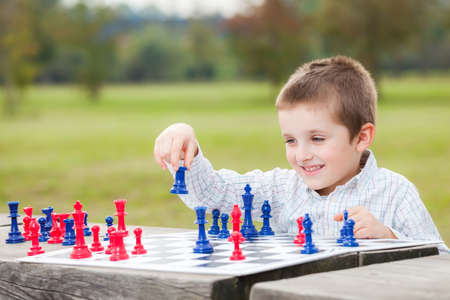 Elegant young boy in white shirt learning to play chess with blue and red chess pieces on wood table in the parkの写真素材