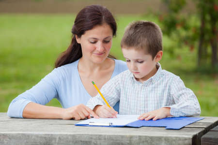 Little young boy in white shirt and his mother write or draw with a pencil on a sheet of paper on wood table in the parkの写真素材