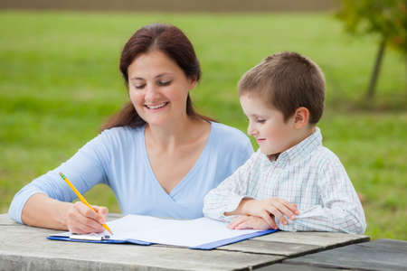 Young woman teacher teaches little young boy in white shirt writing or drawing with a pencil on a sheet of paper on wood table in the parkの写真素材