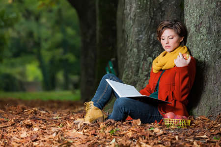 Sexy beautiful serious romantic girl sitting against a tree reading a book eating an apple in the parkの写真素材