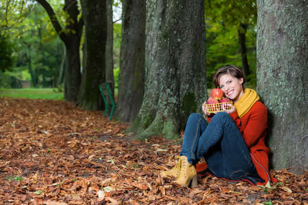Sexy beautiful serious romantic girl sitting against a tree holding a basket with apples in the parkの写真素材