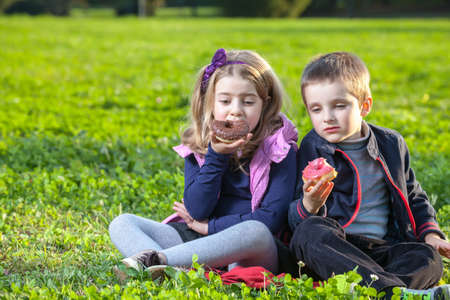 happy kids eating donuts sitting on the green grass in the parkの写真素材
