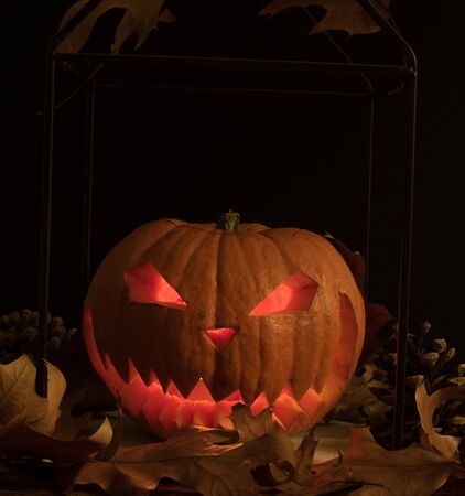 Scary and spooky hand carved Halloween pumpkin with flames inside and dark black background with autumn leaves and cones. Copy and space for text for festive visual representation cardsの写真素材