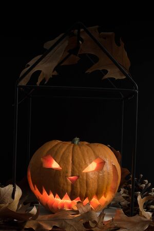Scary and spooky hand carved Halloween pumpkin with flames inside and dark black background with autumn leaves and cones. Copy and space for text for festive visual representation cardsの写真素材