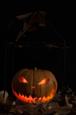 Scary and spooky hand carved Halloween pumpkin with flames inside and dark black background with autumn leaves and cones. Copy and space for text for festive visual representation cardsの写真素材