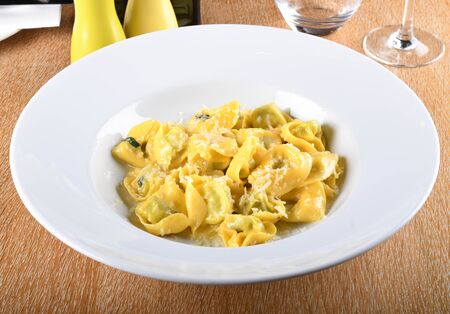 Tortellini with ricotta cheese and spinach, butter, sage sauce, grated parmesan. Parmigiano-Reggiano flakes over traditional italian pasta cooked, served in white plate, over brown table in restaurantの写真素材