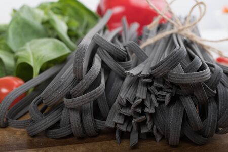 Black squid ink tinted tagliatelle pasta, uncooked, with basil and tomatoes in the background, on a wooden boardの写真素材