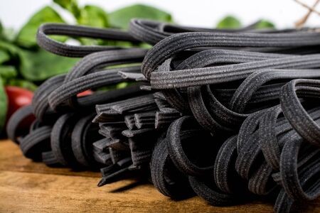 Black squid ink tinted tagliatelle pasta, uncooked, with basil and tomatoes in the background, on a wooden boardの写真素材