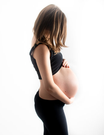 Pregnant woman model touching her pregnancy belly abdomen, isolated on white background. Slim skinny fit blonde lady closeup dressed in black top and leggingsの写真素材