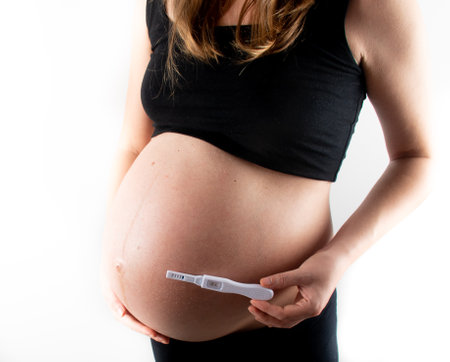 Pregnant skinny slim fit woman holding a positive pregnancy test on her belly tummy abdomen. black top and leggings isolated model on white background concept. new life new beginningsの写真素材