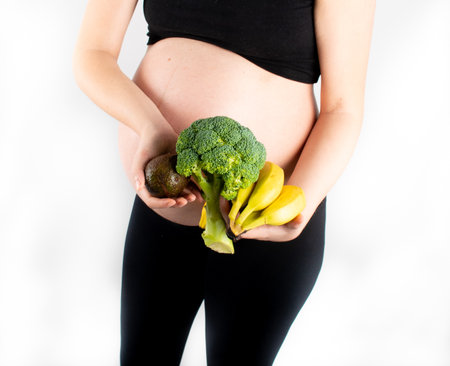 Pregnant woman holding vegetables in hands. Healthy eating concept. Pregnancy, slim skinny girl in black top and leggings isolated on white background. Diet and freshnessの写真素材