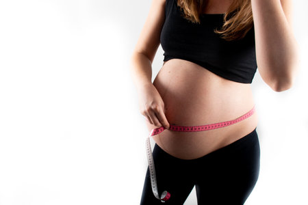 Young pregnant woman measures her waist with measurement tape, weight diet and healthy lifestyle pregnancy concept. Black top and leggings isolated model on white backgroundの写真素材