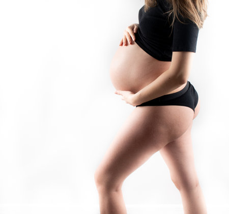 Pregnant woman model touching her pregnancy belly abdomen, isolated on white background. Slim skinny fit blonde lady closeup dressed in black top and leggingsの写真素材