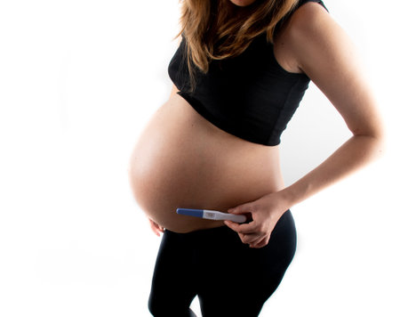 Pregnant skinny slim fit woman holding a positive pregnancy test on her belly tummy abdomen. black top and leggings isolated model on white background concept. new life new beginningsの写真素材