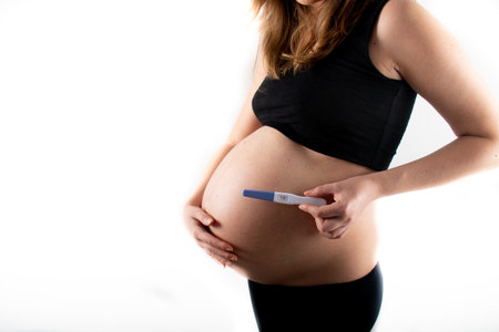 Pregnant skinny slim fit woman holding a positive pregnancy test on her belly tummy abdomen. black top and leggings isolated model on white background concept. new life new beginningsの写真素材