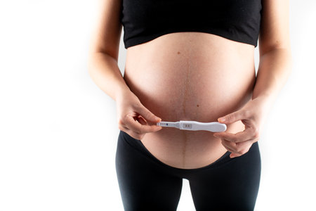 Pregnant skinny slim fit woman holding a positive pregnancy test on her belly tummy abdomen. black top and leggings isolated model on white background concept. new life new beginningsの写真素材
