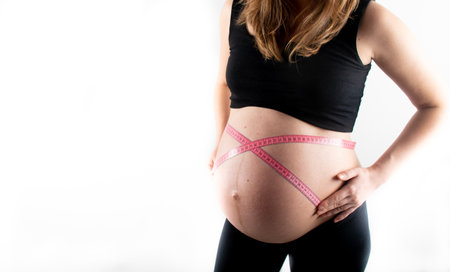 Young pregnant woman measures her waist with measurement tape, weight diet and healthy lifestyle pregnancy concept. Black top and leggings isolated model on white backgroundの写真素材