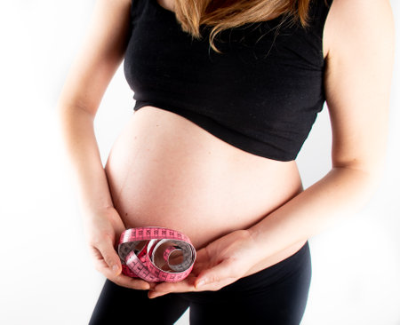 Young pregnant woman measures her waist with measurement tape, weight diet and healthy lifestyle pregnancy concept. Black top and leggings isolated model on white backgroundの写真素材